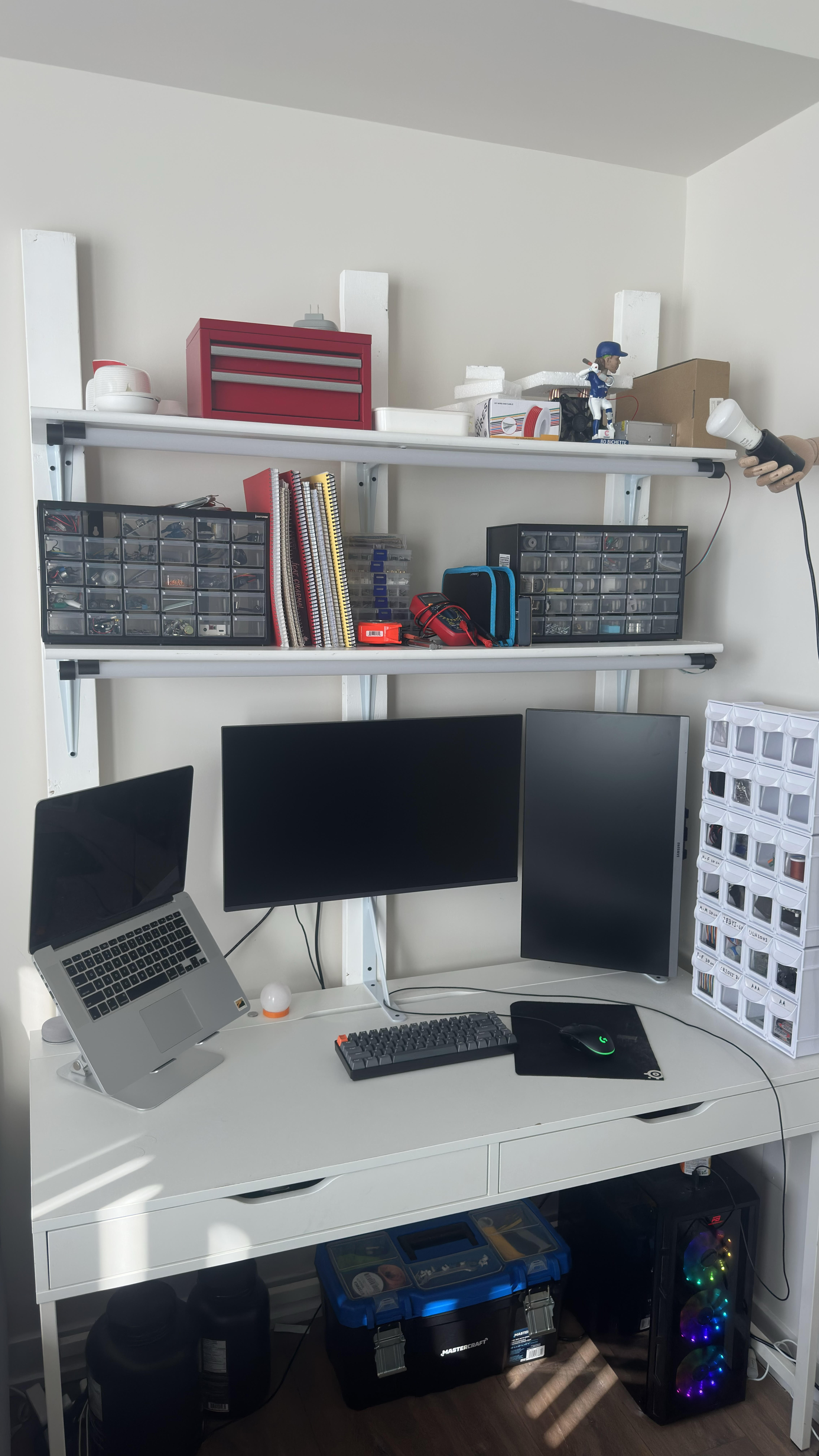 Desk setup: IKEA Alex with 2×4 uprights, white shelves with organizers and toolbox, dual monitors and laptop on the desktop, PC tower below