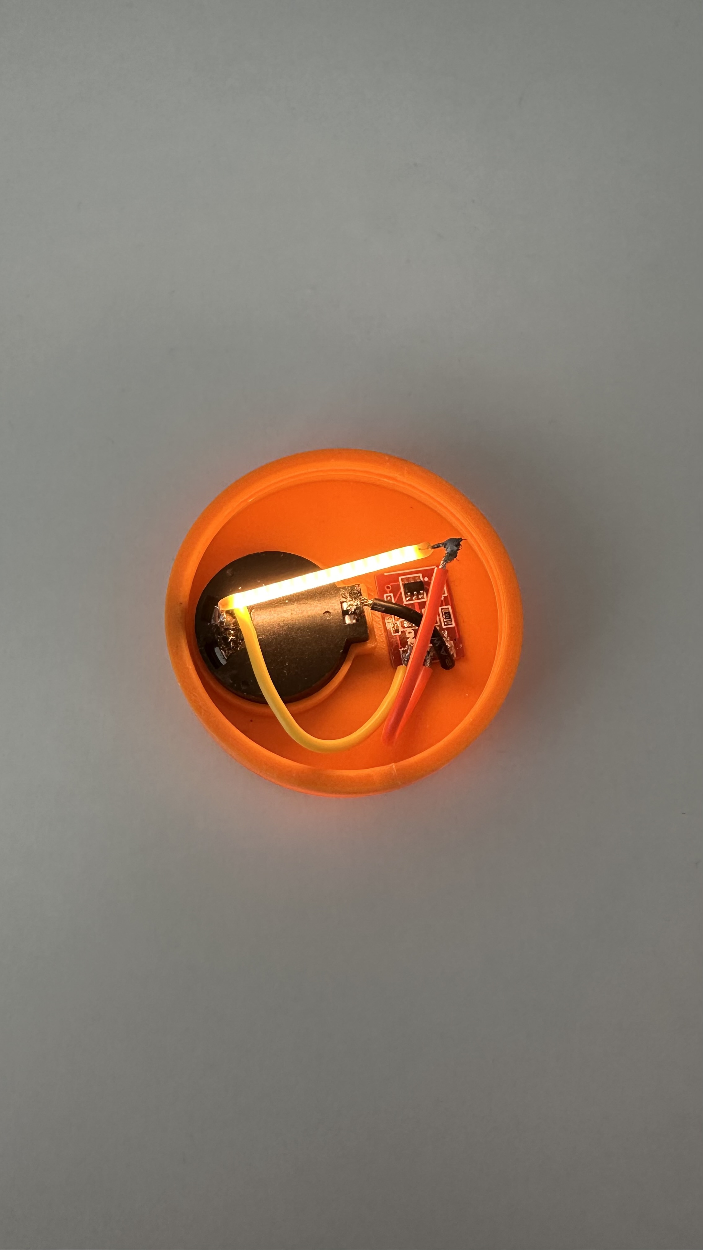 Top view into orange base: coin cell, green TTP223 board, and glowing LED filament