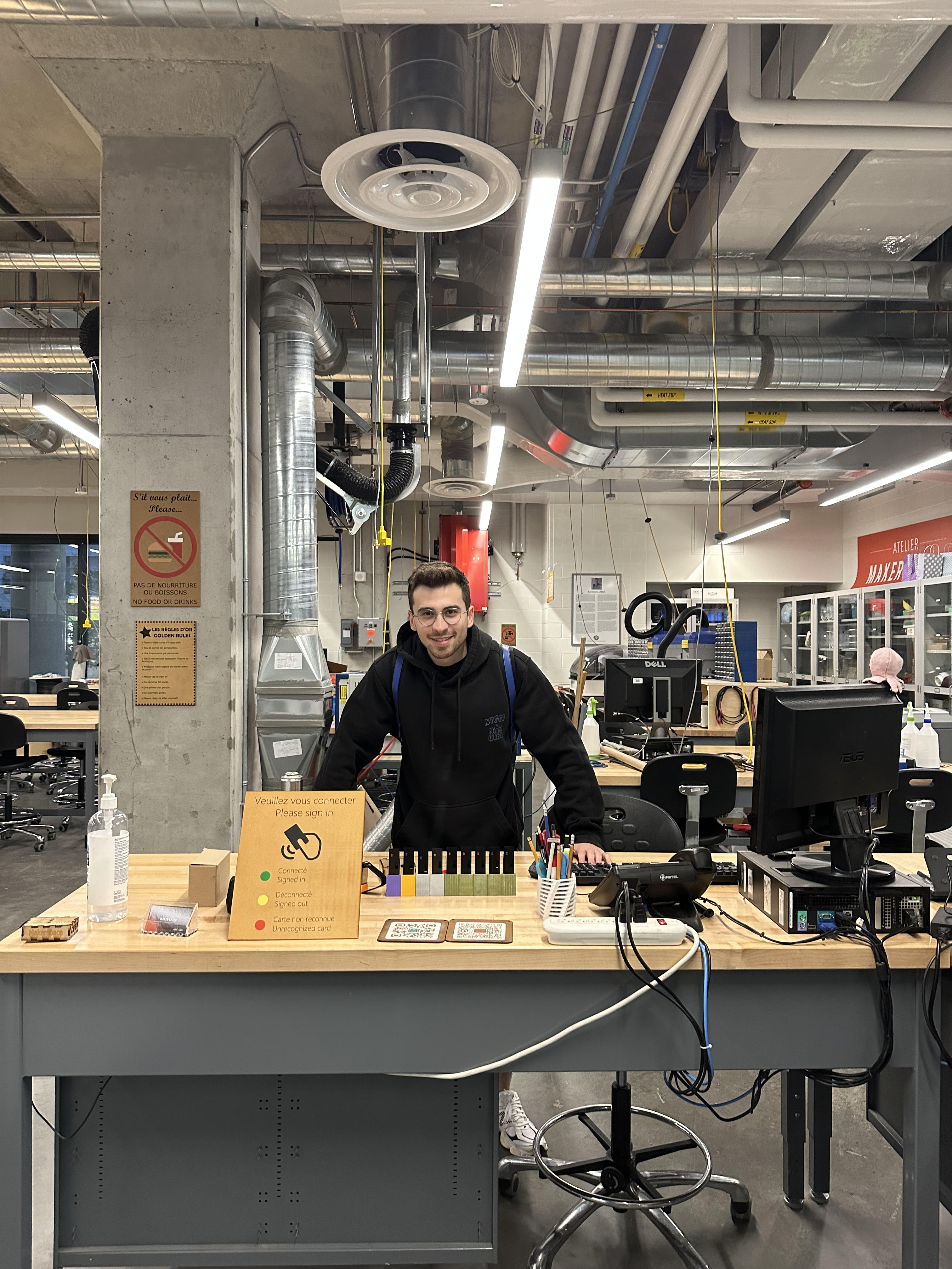 Makerspace workbench: staff behind a wooden bench with monitor and tools, industrial ceiling and lighting